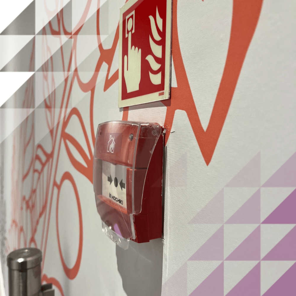 Un pulsante manuale di allarme antincendio Hochiki rosso con copertura protettiva, montato sotto un cartello indicante la presenza di un pulsante di allarme antincendio su una parete bianca decorata con un murale grafico di colore arancione acceso.