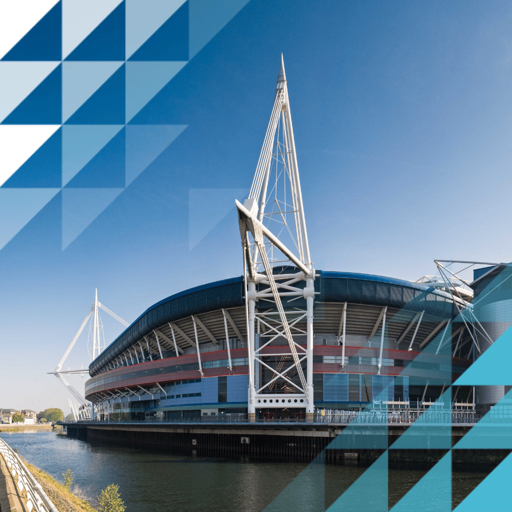 Exterior view of the Principality Stadium in Cardiff, seen from the riverside on a clear sunny day, with its distinctive white steel masts and retractable roof structure visible against a blue sky.