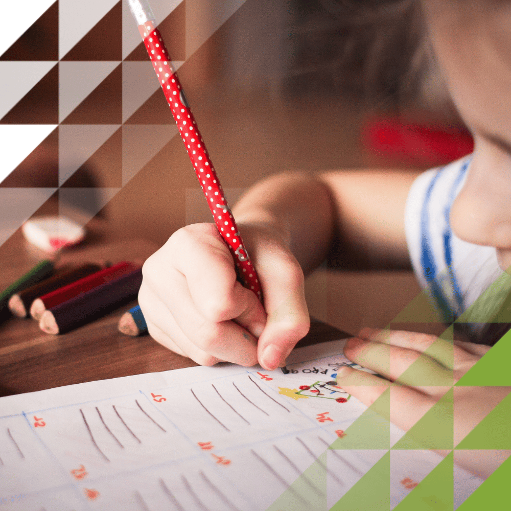 Close-up of a child's hand writing with a red polka-dot pencil on a lined worksheet with numbered rows and drawings, with colored pencils on the table nearby.