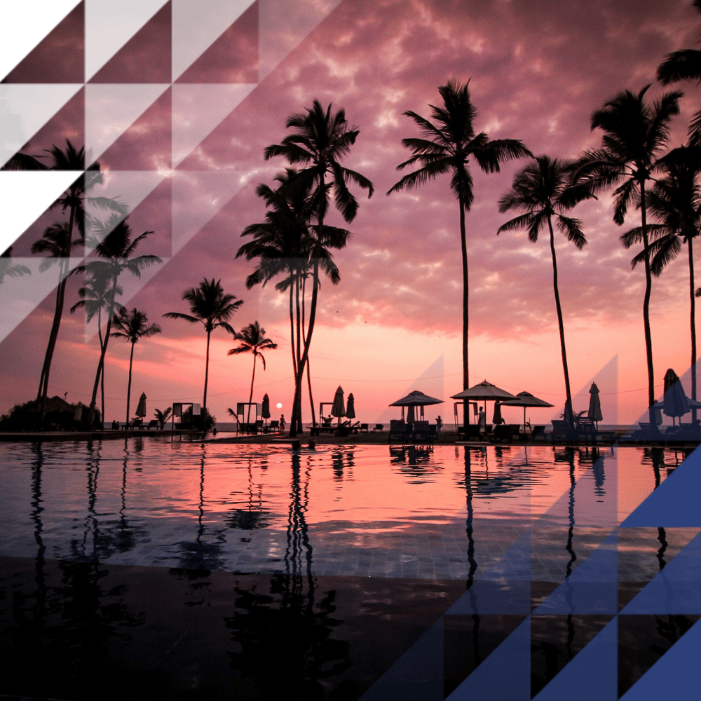 A luxury hotel infinity pool at sunset, with silhouetted palm trees and parasols reflected in the still water against a dramatic pink and purple sky.