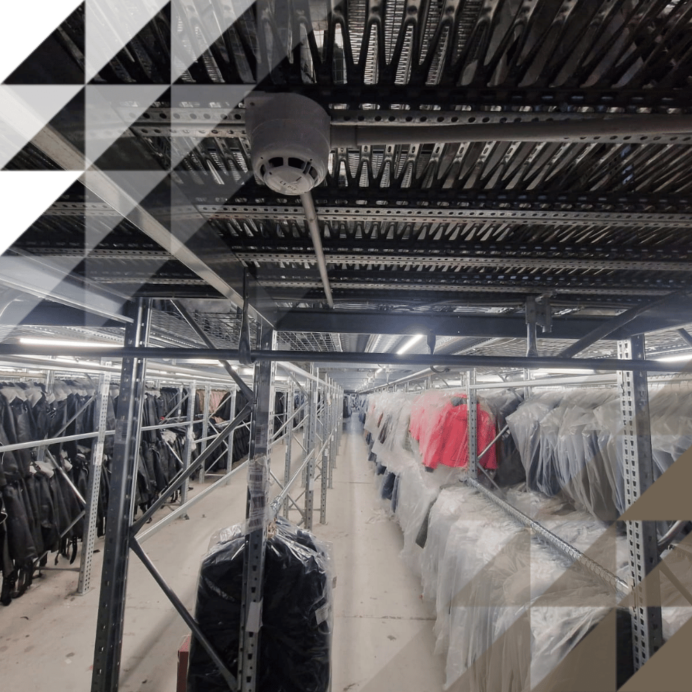 Narrow corridor between tall metal racking units densely packed with wrapped hanging garments, with strip lighting and smoke detector overhead.
