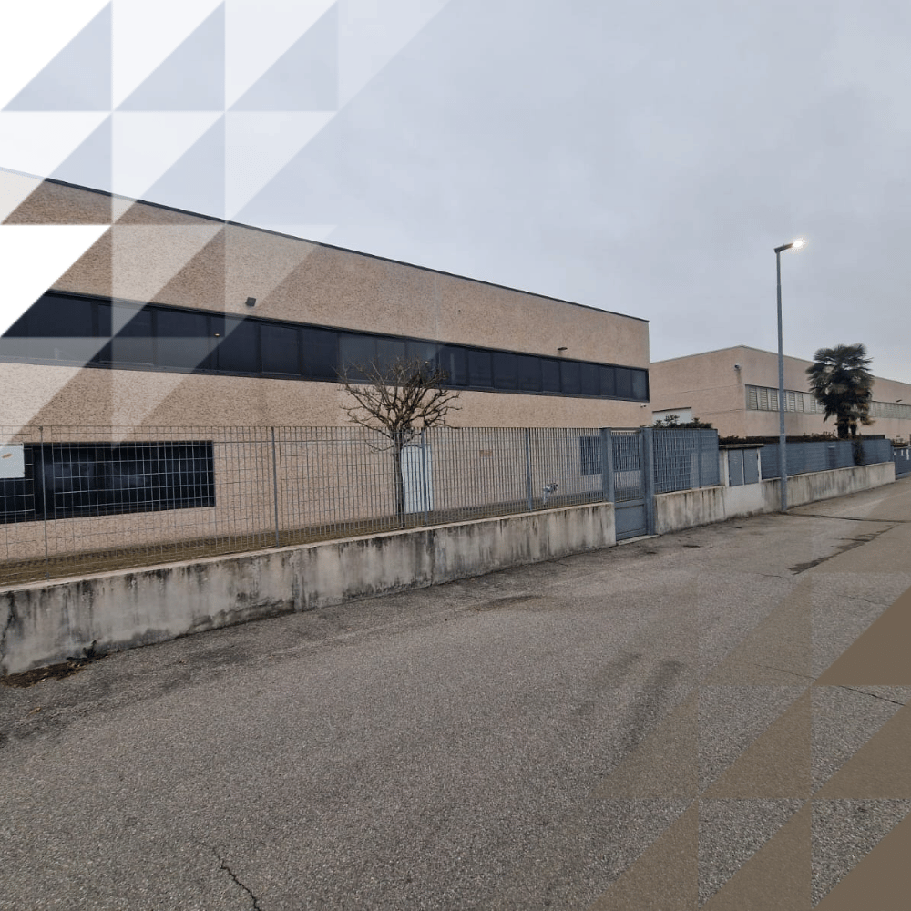 Street-level view of a two-storey warehouse building with beige brick exterior, strip windows and metal security fence, under an overcast sky.