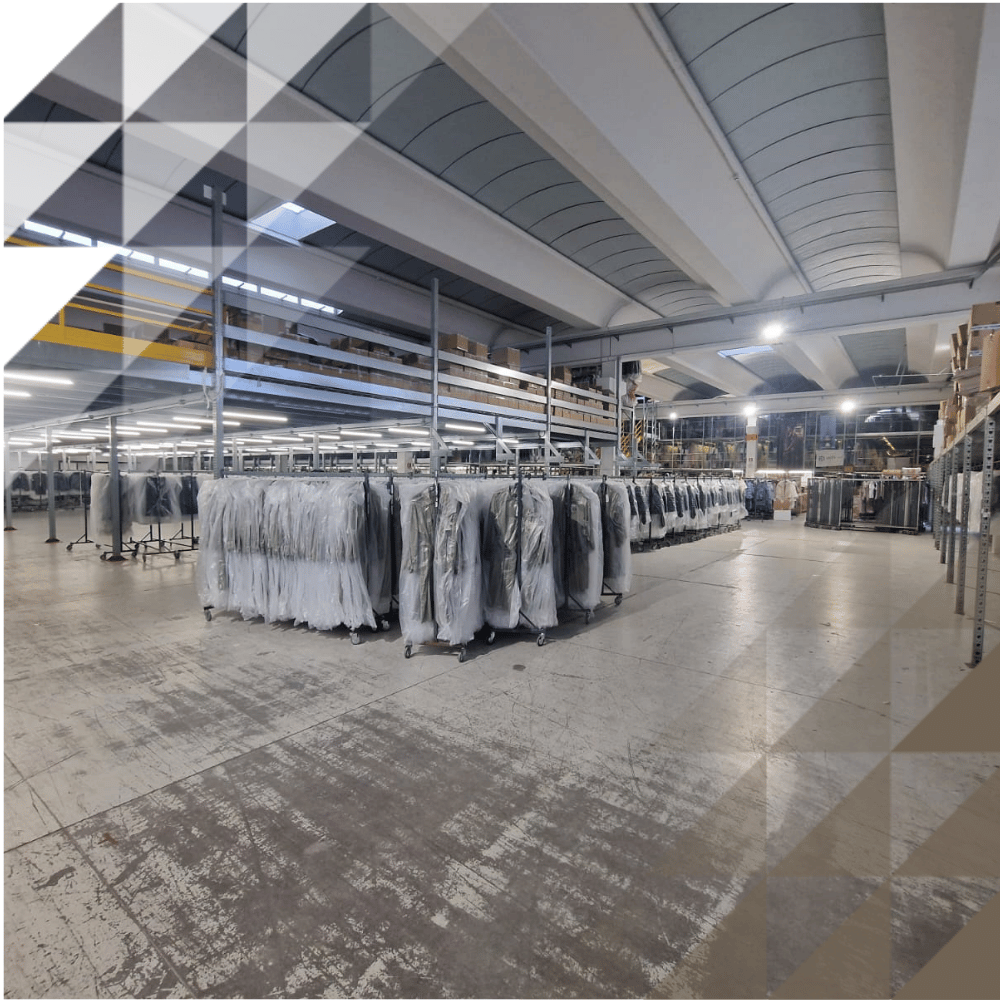 Wide-angle view of large warehouse with exposed steel ceiling, fluorescent strip lighting and multiple rows of garments on hanging rails.