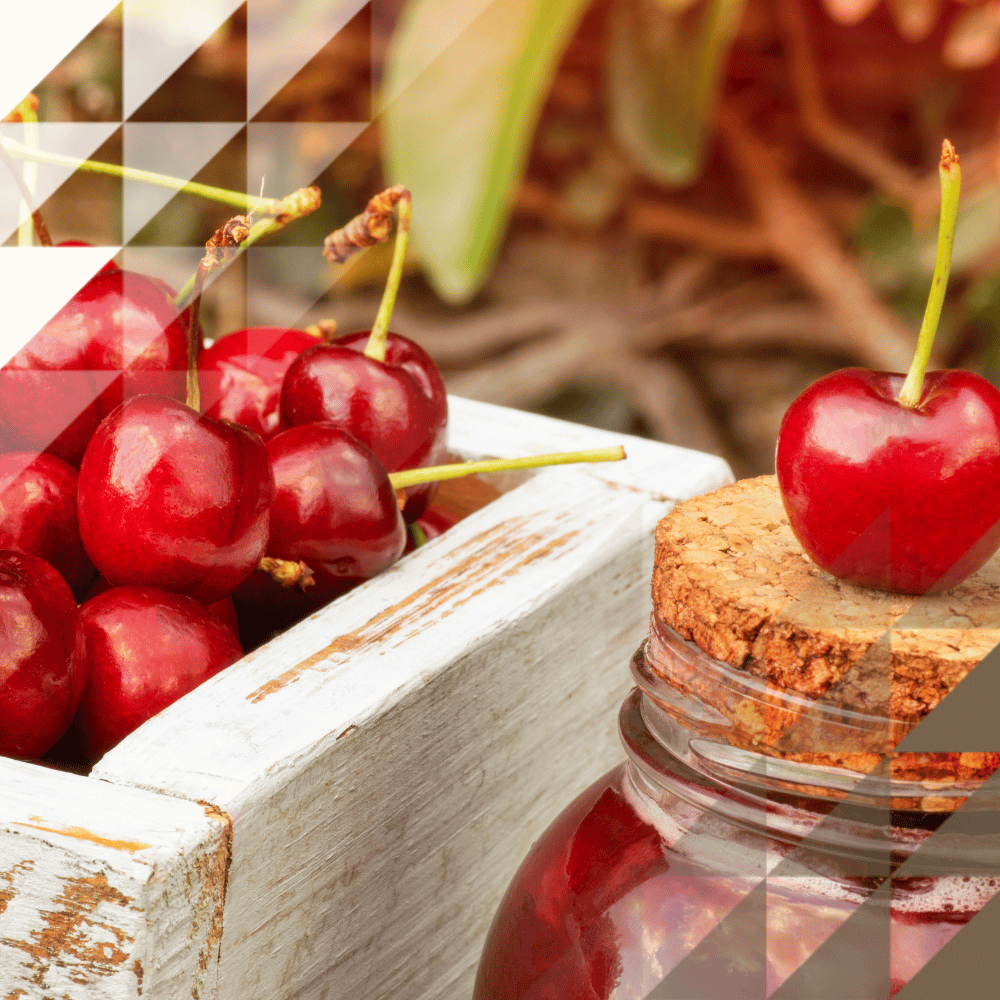 A close-up of bright red cherries with stalks heaped in a distressed white wooden box, with a small cork-lidded glass jar topped with a single cherry placed beside it, against a blurred natural background.