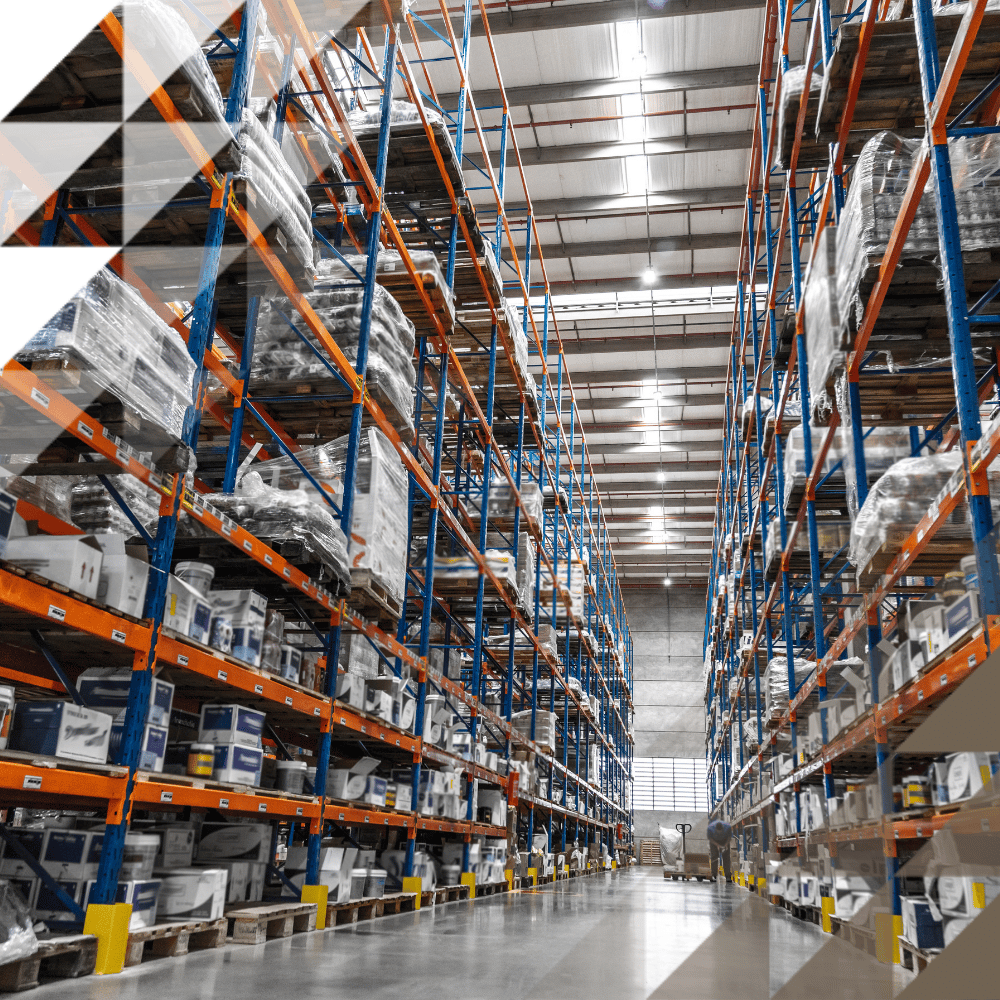 Una vista dal livello del suolo lungo una lunga corsia di magazzino, fiancheggiata da imponenti scaffalature per pallet blu e arancioni, densamente riempite di merci in scatole e pallet avvolti in pellicola, illuminate da luci industriali a soffitto, con un operaio visibile in lontananza.