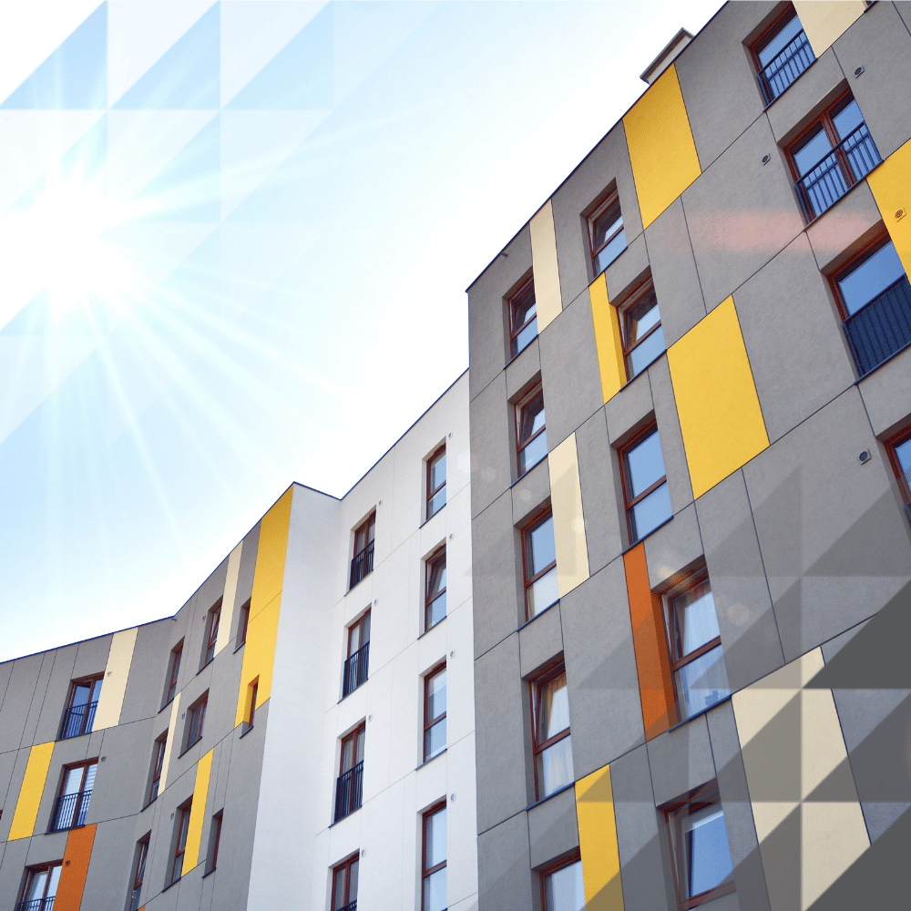 Low-angle view of a modern residential apartment block with a colourful grey, yellow, orange, and cream cladded facade, photographed against a bright sunny sky with lens flare.