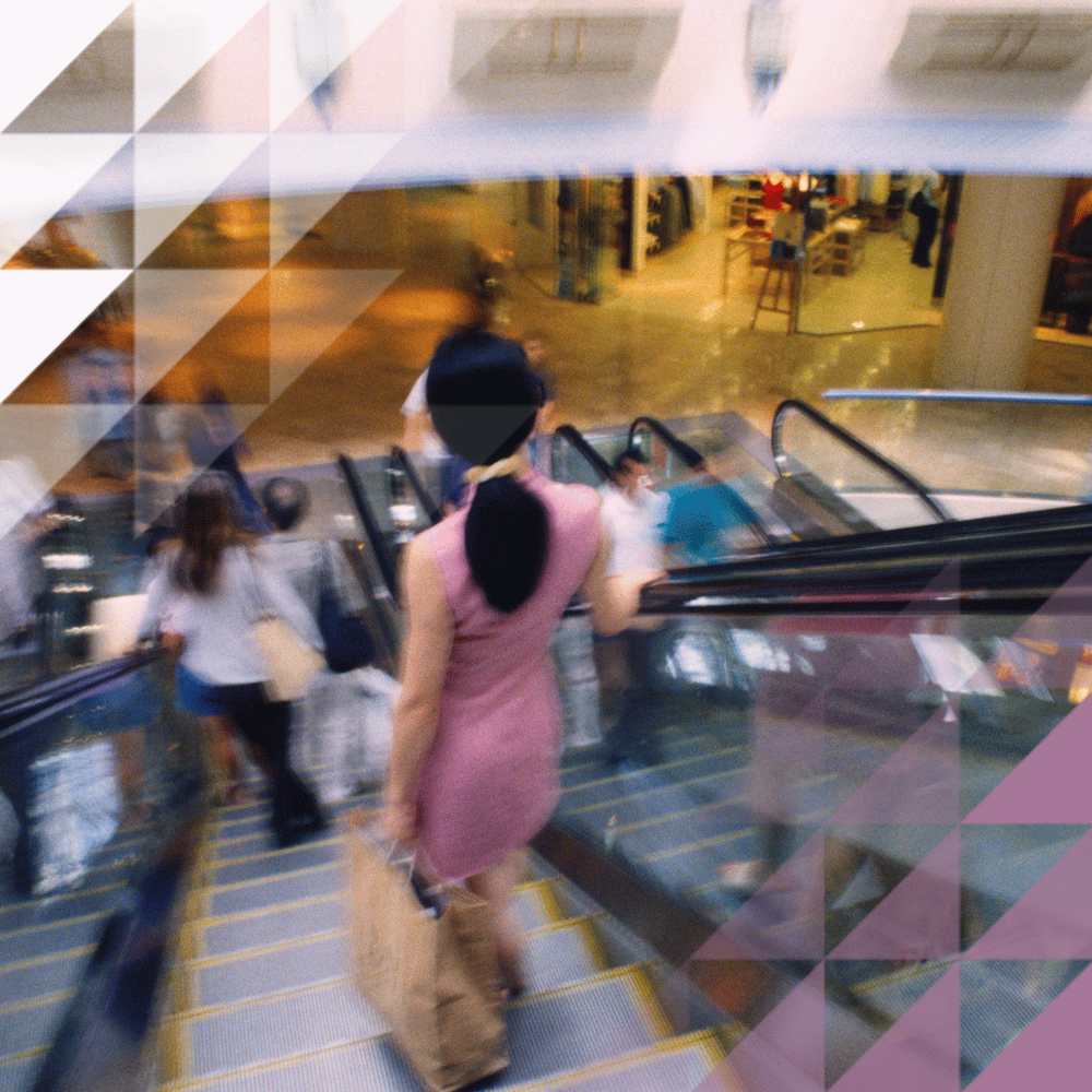Motion-blurred shoppers riding escalators in a busy multi-level shopping mall, with retail units visible on the floor below.