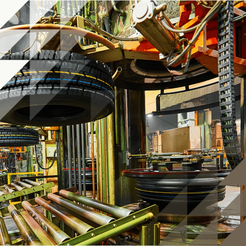 Freshly moulded tyres moving along an automated conveyor system inside a large industrial factory, with heavy green machinery and equipment visible throughout.