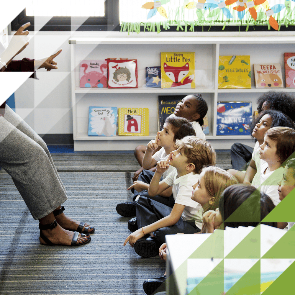 Un’insegnante sorridente che parla a dei bambini piccoli seduti sul pavimento in un’aula di scuola elementare decorata con colori vivaci.
