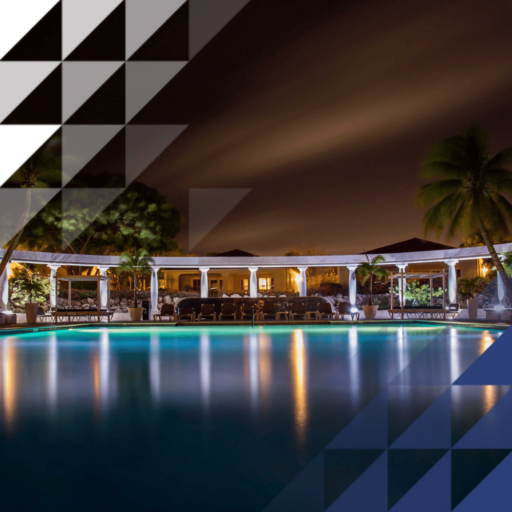 Nighttime view of a luxury resort pool illuminated with warm lights, framed by white colonnades, palm trees, and poolside loungers, with the lights reflecting on the calm turquoise water under a dark sky.