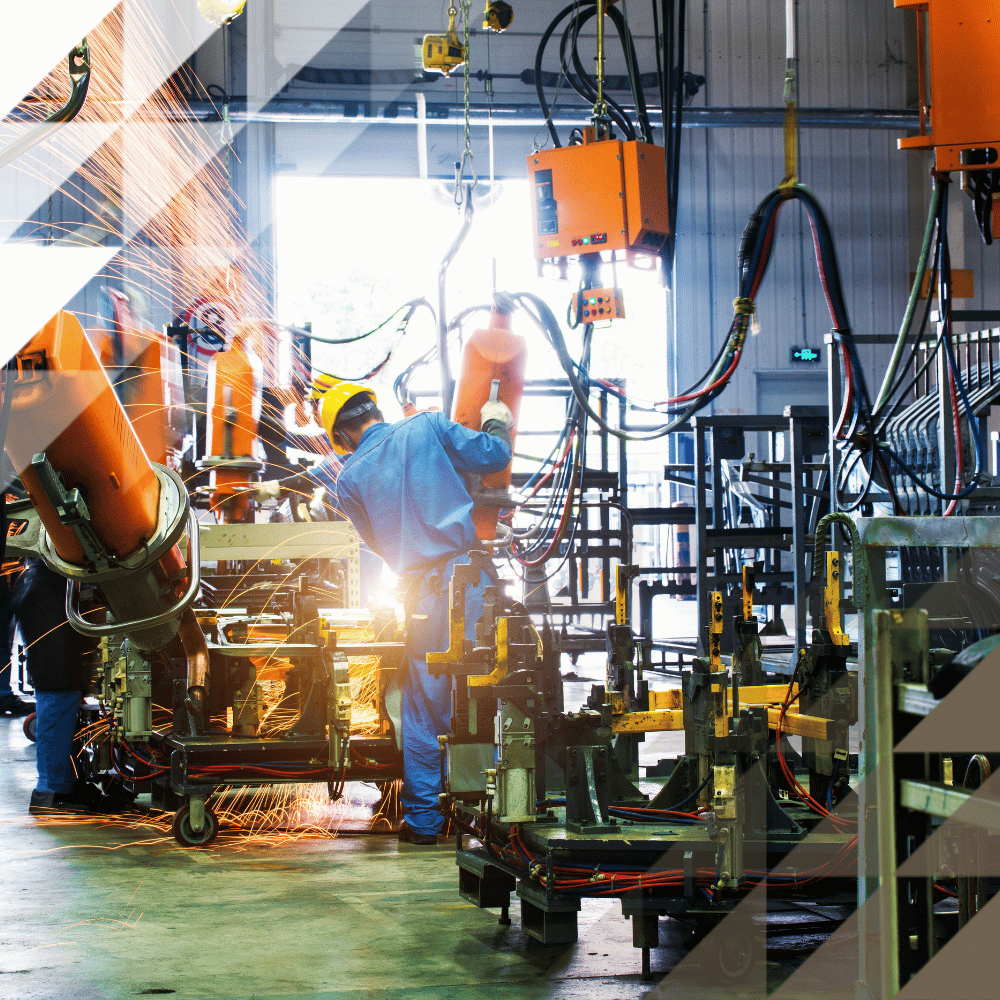 A worker in blue coveralls and a hard hat welds metal parts on a factory production line, surrounded by orange robotic arms, sparks, and manufacturing equipment.