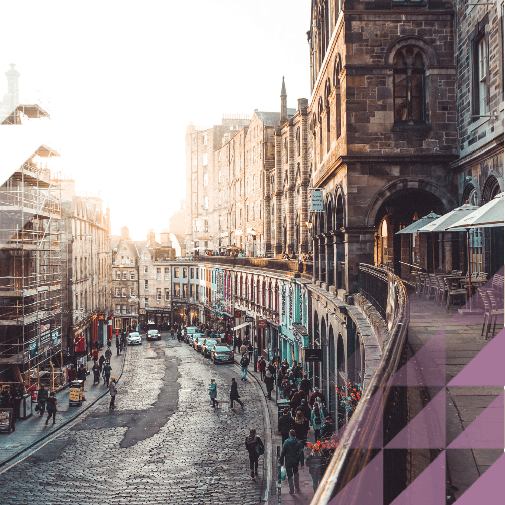 High-angle view of busy cobbled Edinburgh Old Town street, with colourful shops, pedestrians, and a café terrace with parasols beside a stone building.
