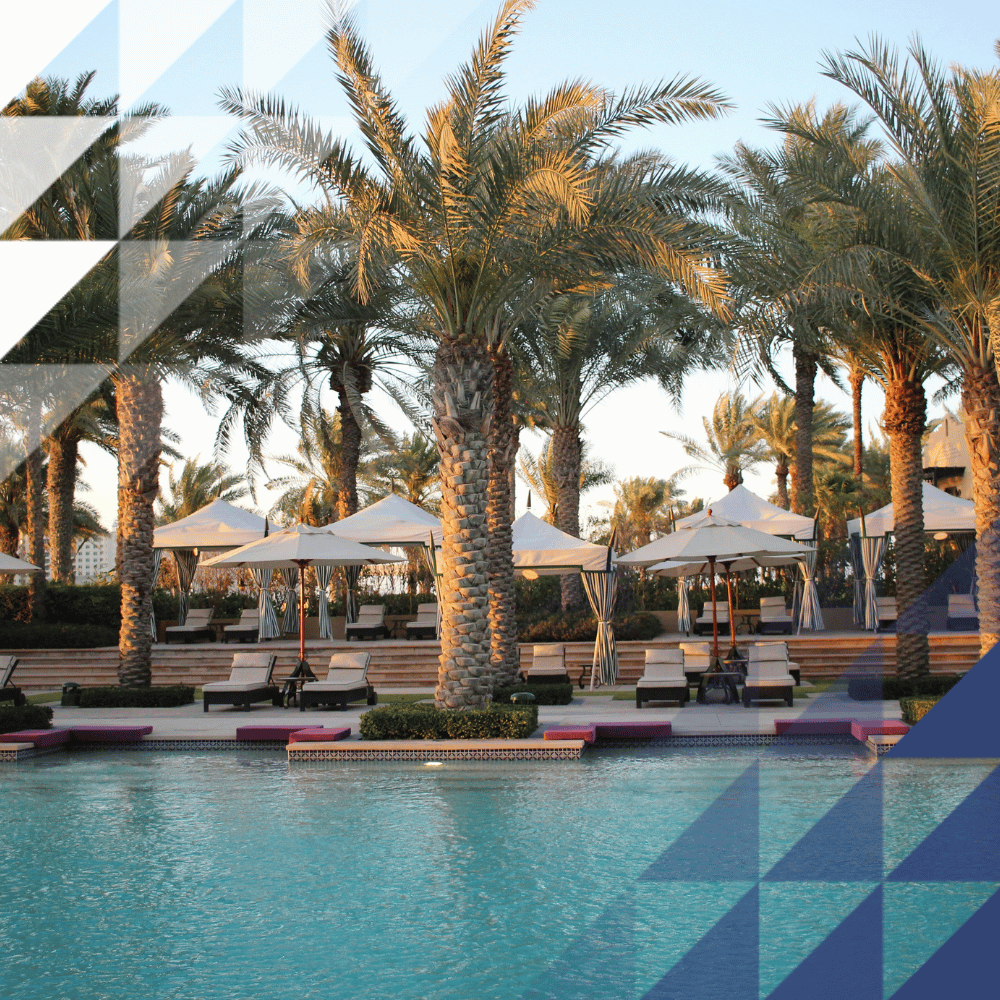 Turquoise pool bordered by date palms, with sun loungers, white parasols and pink poolside platforms in warm evening light.