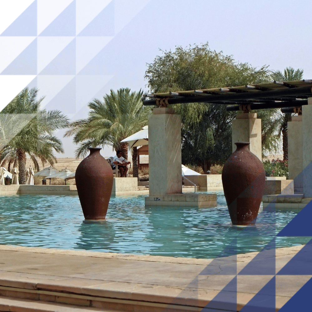 Resort pool with terracotta urns, stone colonnades and timber pergola, surrounded by date palms and desert landscape.