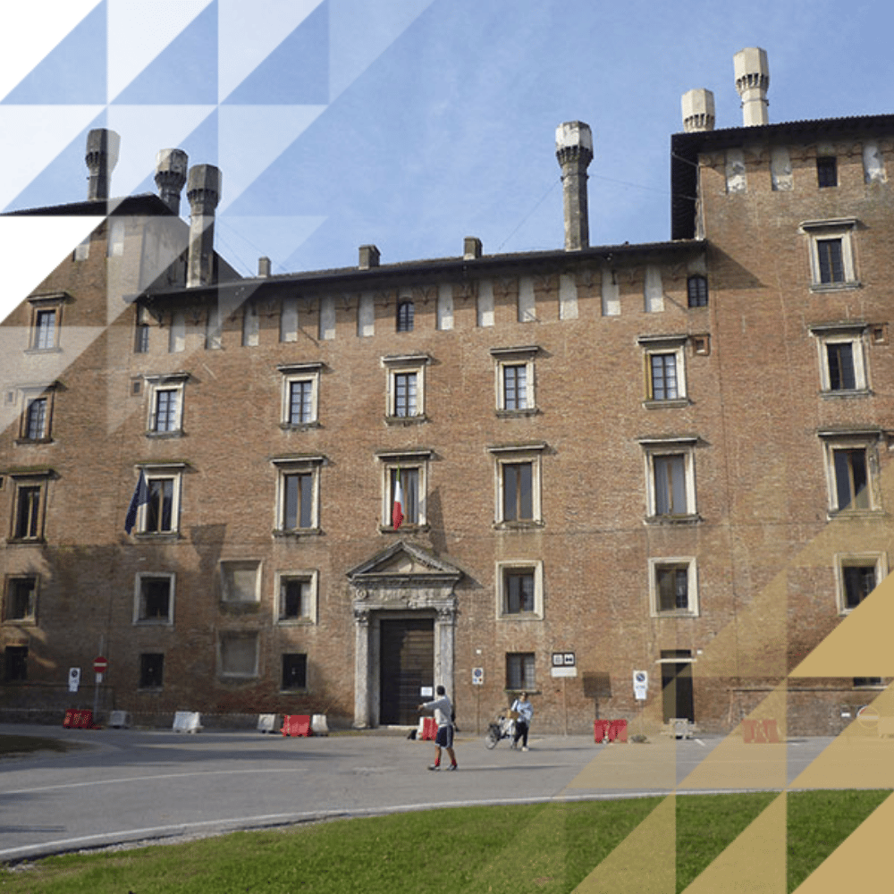 Historic multi-story Italian brick palazzo with tall chimney stacks, arched windows, and a central ornate entrance, with Italian and EU flags and a few people in the courtyard below.