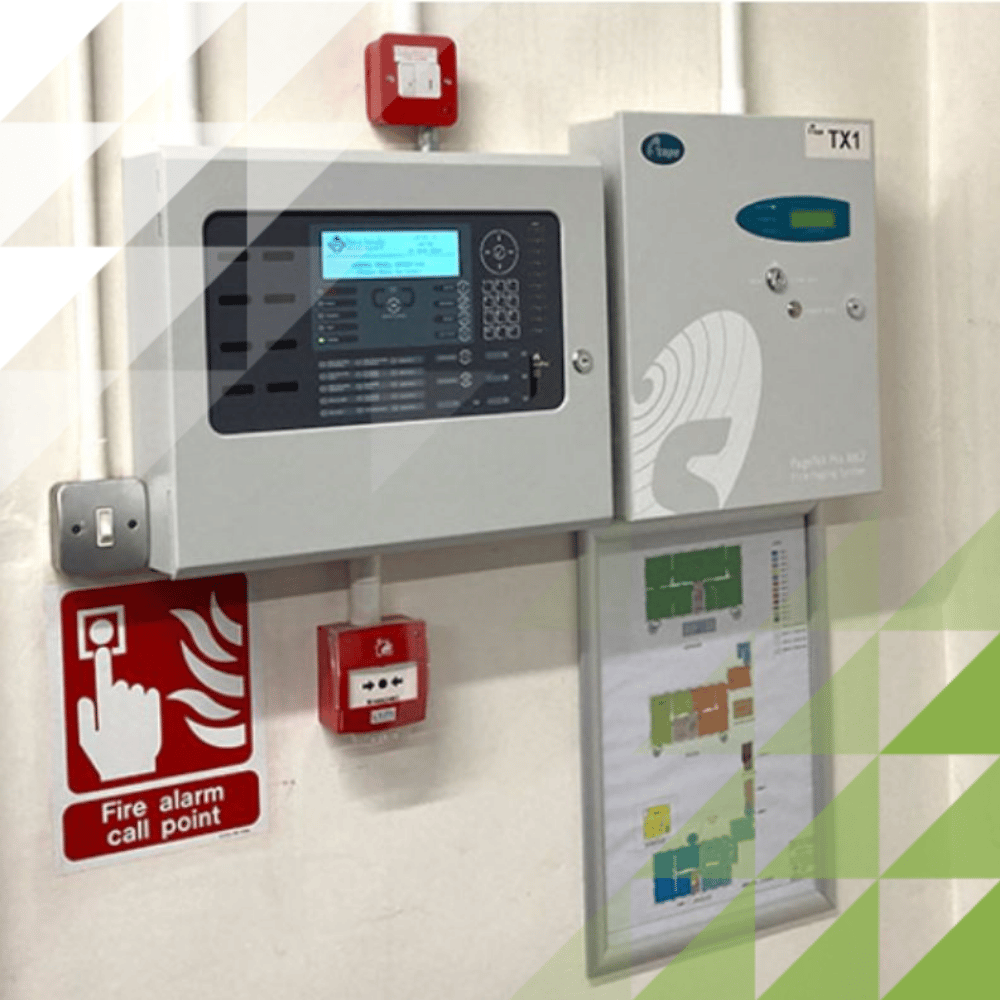Wall-mounted addressable fire alarm control panel with LCD display, manual call point, TX1 ancillary unit and zoned floor plan chart.