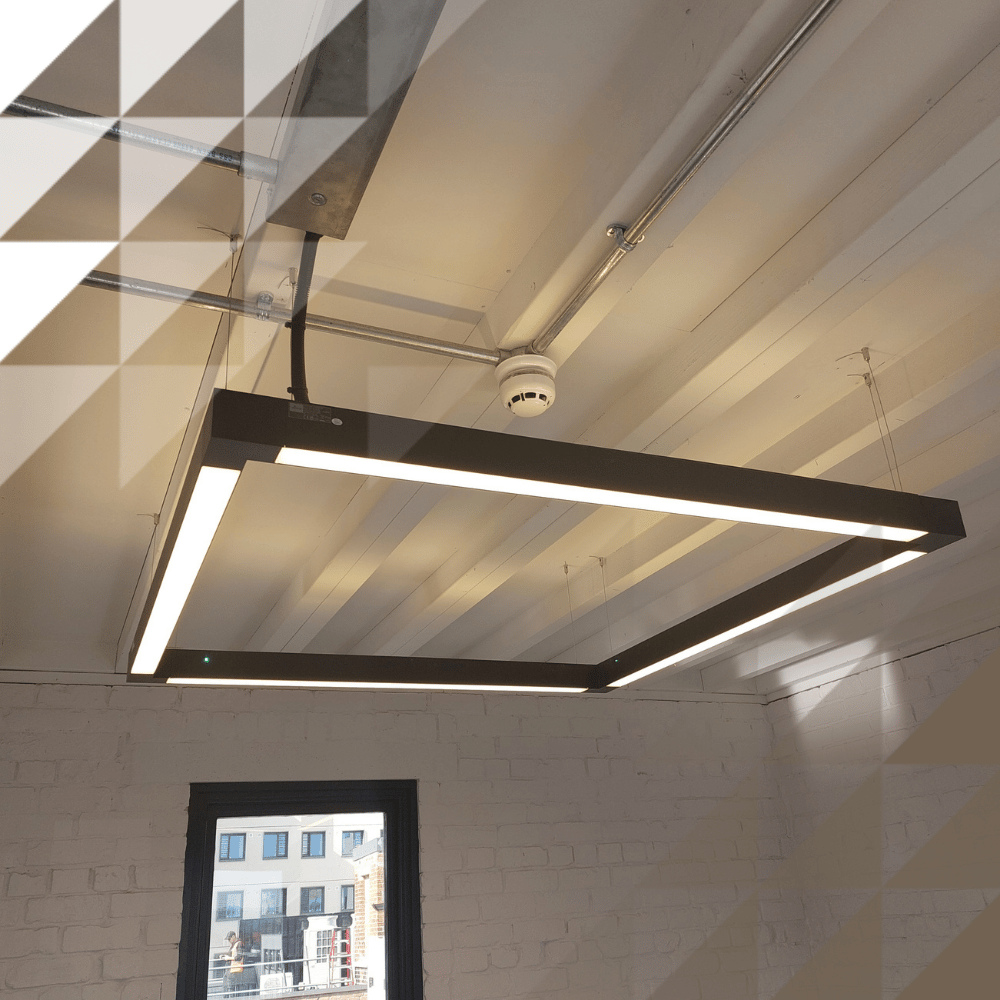 Upward view of a square black LED light fixture suspended from a white-painted ceiling with exposed pipes and a smoke detector, in a room with white-painted brick walls and a dark-framed window overlooking nearby buildings.