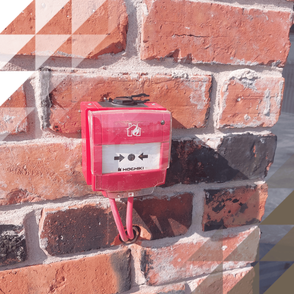 Close-up of a red Hochiki manual fire alarm call point mounted on an exposed red brick wall, with a push-arrows label and protective cover.