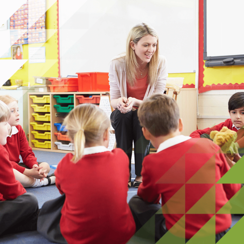 A smiling teacher sits on a chair talking with young pupils in red school uniforms seated on the classroom floor around her, with colourful storage bins and a whiteboard in the background.