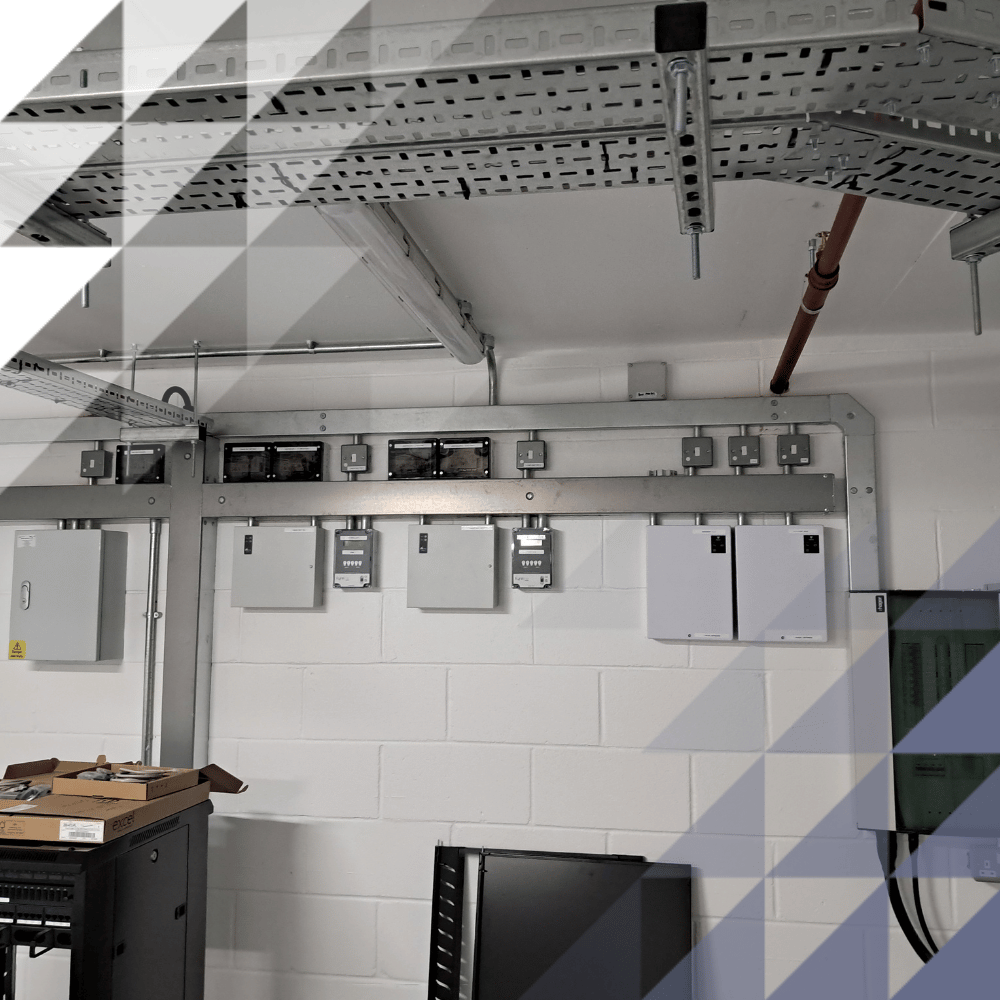 Plant room interior with grey electrical control units and junction boxes on a white brick wall, connected by overhead cable trays and conduit.