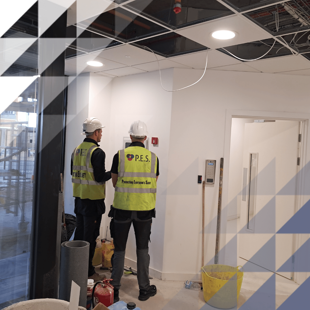 Two P.E.S. workers in hi-vis vests and hard hats examining a fire alarm panel inside a building under construction.
