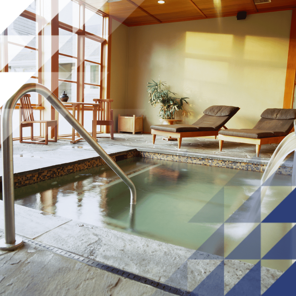 Indoor spa pool with mosaic tile surround and stainless steel handrail, flanked by two cushioned sun loungers, wooden chairs, and a potted plant beside large windows.