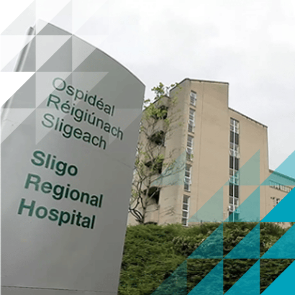 In primo piano, l'insegna dell'Ospedale regionale di Sligo in irlandese e in inglese, con l'edificio a più piani dell'ospedale visibile sullo sfondo.