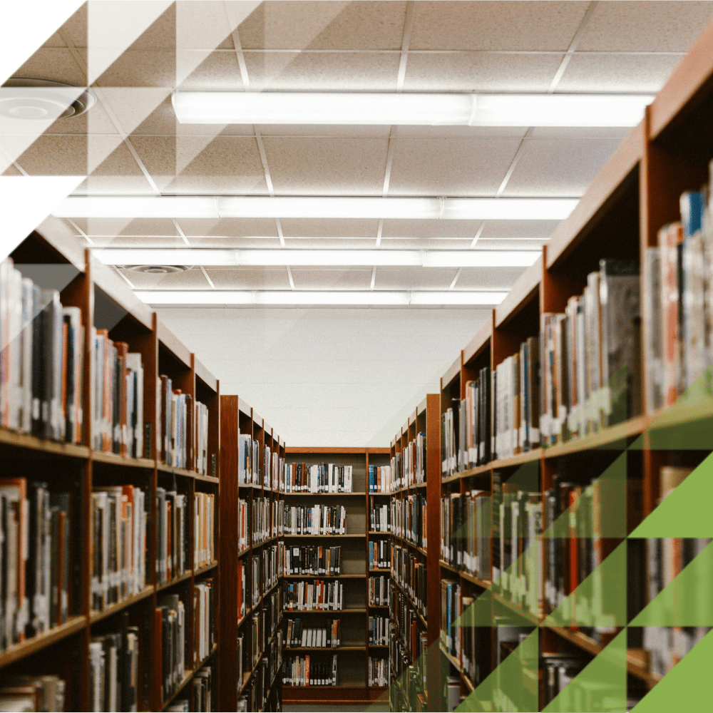Library aisle lined with tall wooden bookshelves, with fluorescent strip lighting and a smoke detector on the ceiling.