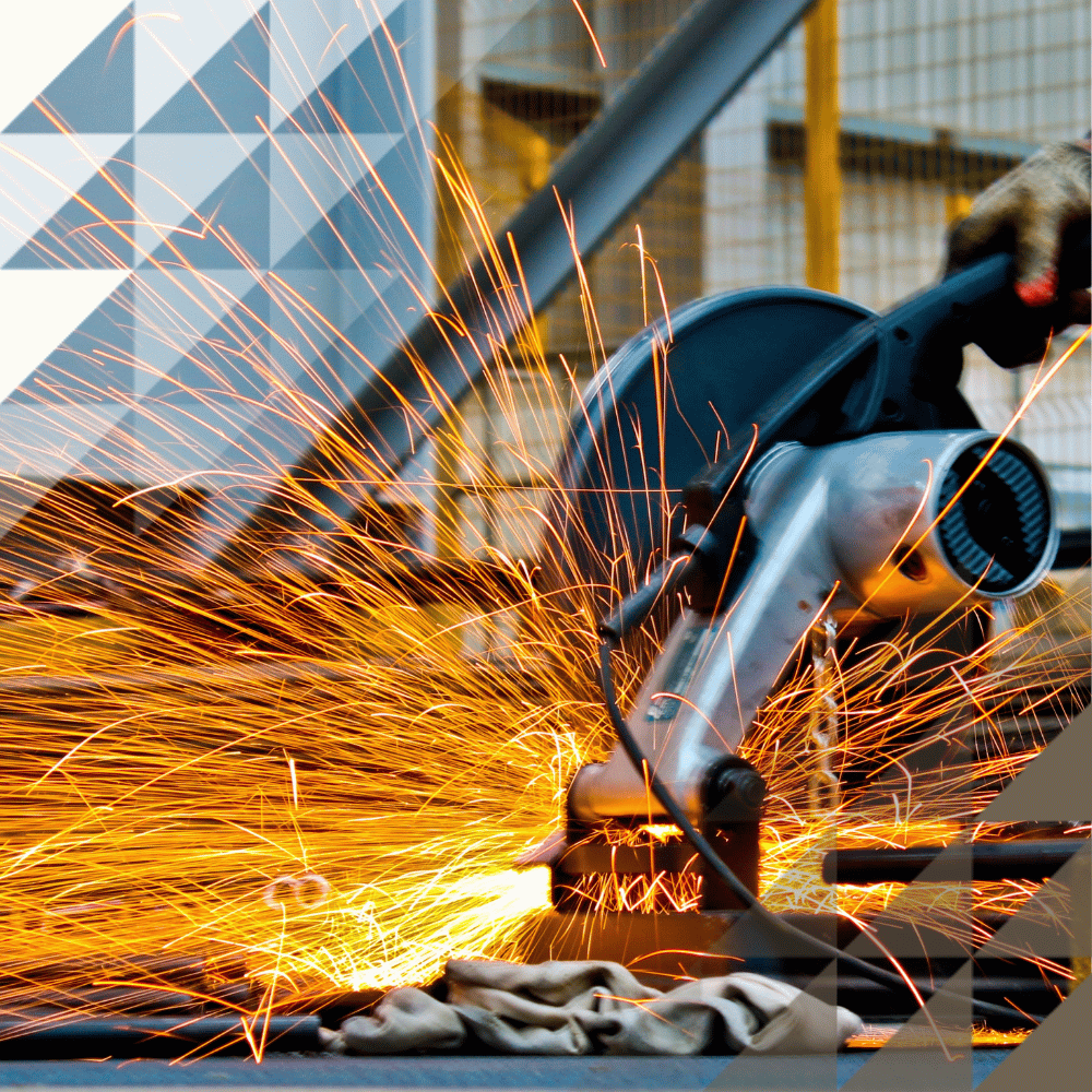 A gloved worker uses an angle grinder on metal at an industrial site, sending a dramatic shower of bright orange sparks across the foreground.