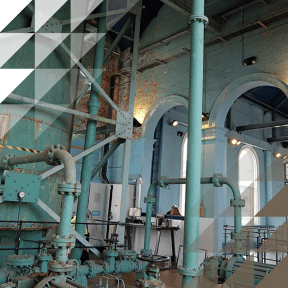 Large teal-painted pipes, valves and structural columns filling the interior of a grand Victorian building with arched windows and exposed brick walls.