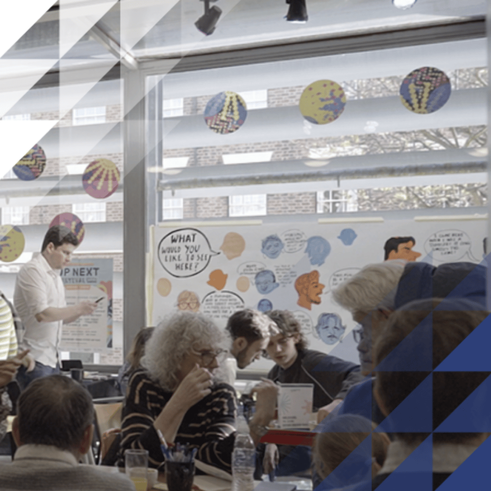 People seated at tables in discussion during a community workshop, with illustrated posters and colourful window decorations, including one reading 