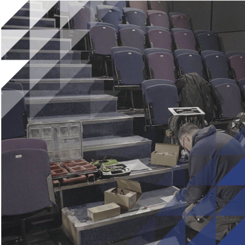 A technician kneels on carpeted theatre steps installing equipment amid open toolboxes and cables, with rows of empty blue and purple auditorium seating in the background.