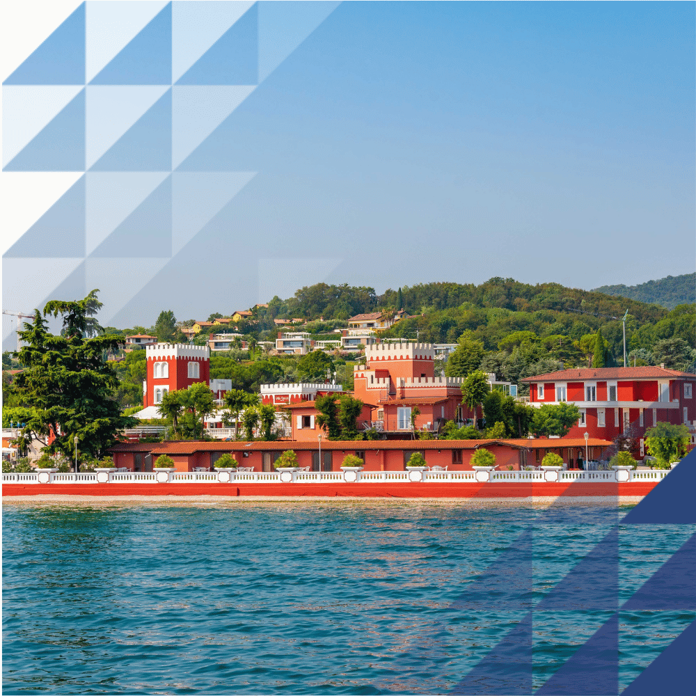 Pastel waterfront buildings above turquoise Lake Garda, with dramatic mountain scenery under a partly cloudy sky.