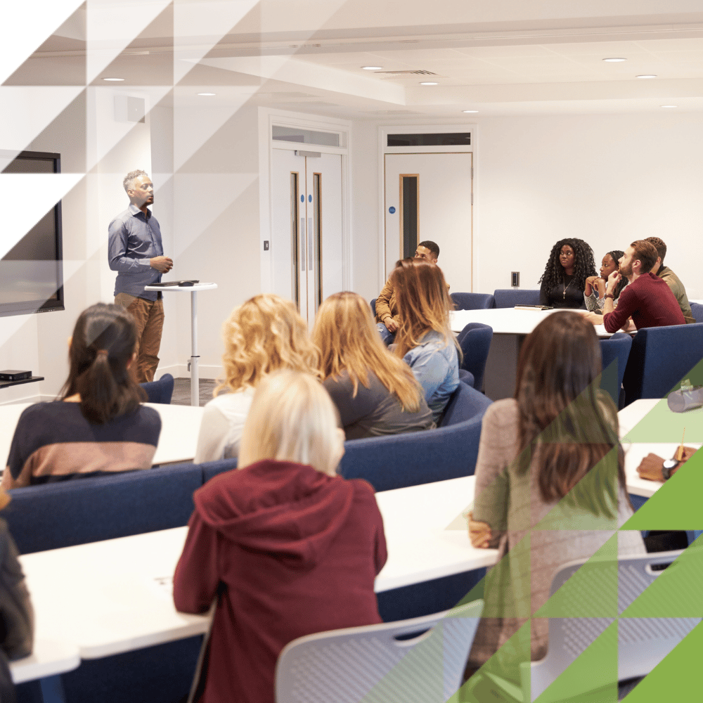 A lecturer addresses a group of students seated at modern collaborative tables in a bright university classroom, with a digital display screen mounted on the wall.