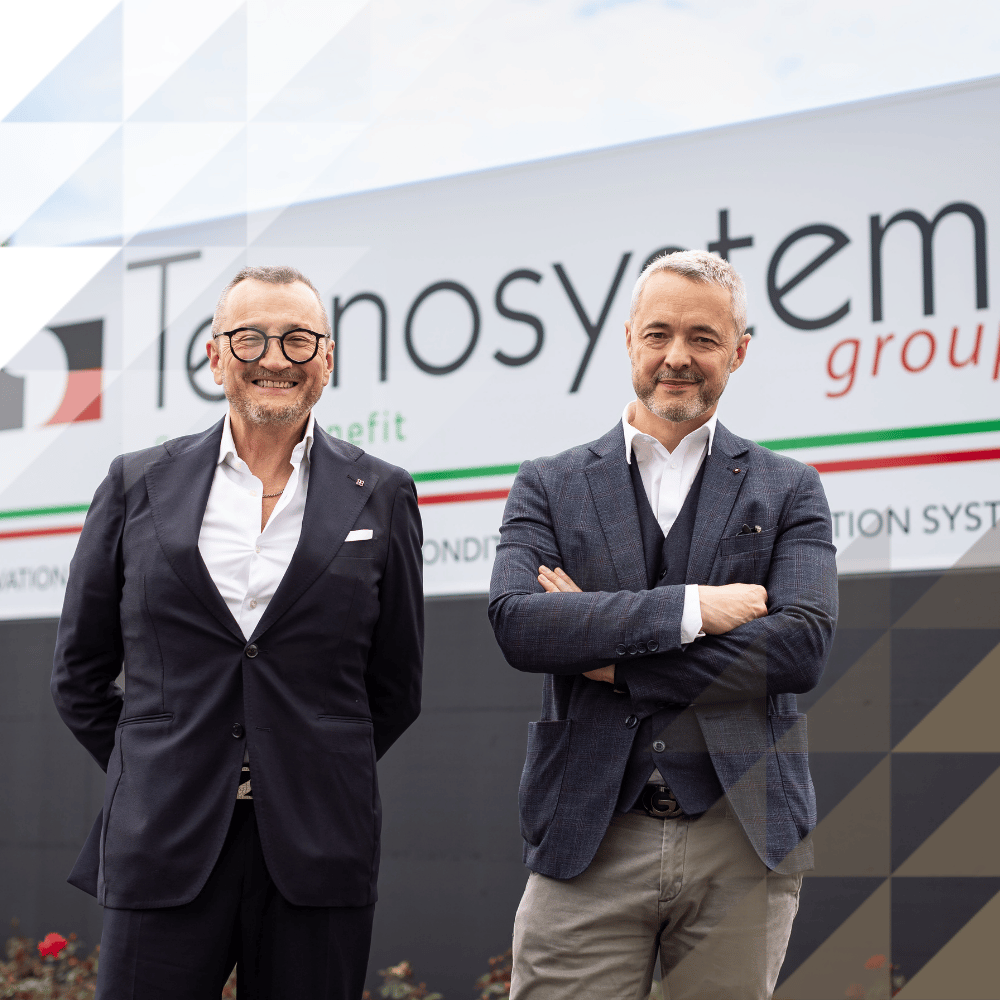 Two smartly dressed men smiling in front of a Tecnosystemi Group branded sign reading 
